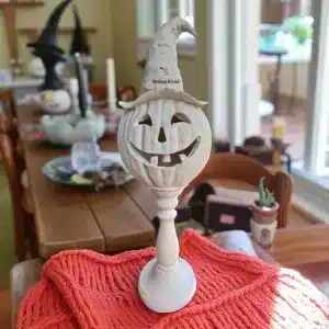 Turuncu renkli örgü bir örtü üzerinde, aydınlık bir ortamda sergilenen, cadı şapkalı ham polyester kabak heykeli (POL78). Halloween masası için dekoratif heykel.